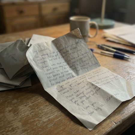 Elegant handwritten letter on textured paper with pens, coffee mug, and stacks of paper on a wooden desk, showcasing professional graphic design services and branding solutions by Alan Wellburn.