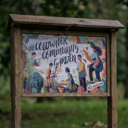Colourful wood-framed sign for Coldwater Community Garden with hand-painted illustrations of people gardening and the words "Coldwater Community Garden" in bold lettering.
